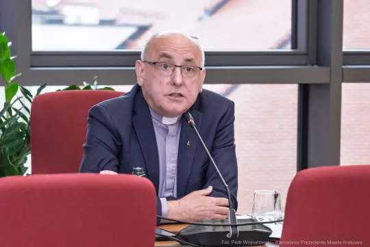 Mężczyzna w okularach, ubrany w garnitur, przemawia podczas spotkania.