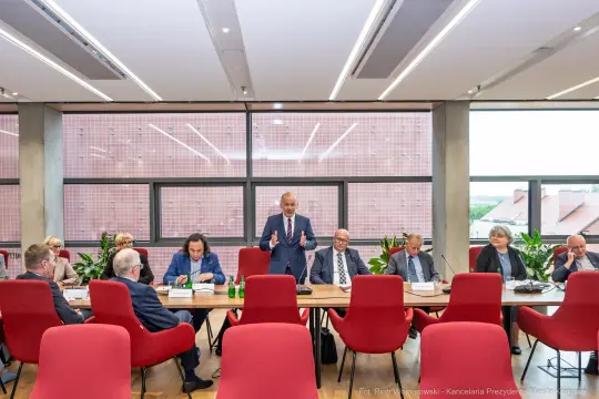 Spotkanie grupy ludzi przy stole, mężczyzna przemawia, w tle okna i rośliny.