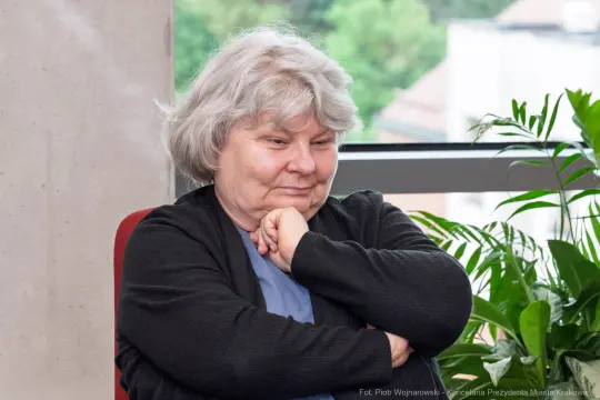 Kobieta z siwymi włosami, zamyślona, siedzi w fotelu obok roślin.