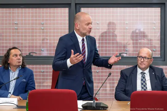 Mężczyzna w garniturze przemawia podczas spotkania, dwaj inni mężczyźni słuchają.