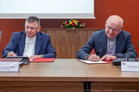 Dwaj mężczyźni podpisują dokumenty przy stole, w tle kwiaty.