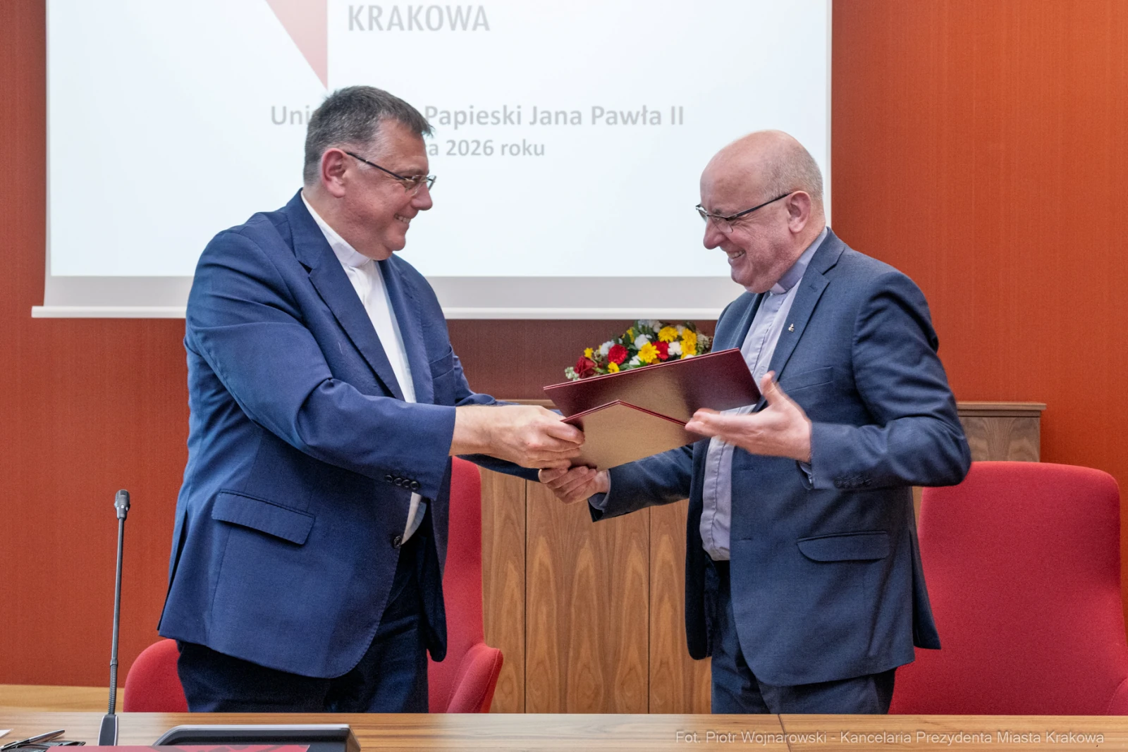 Dwaj mężczyźni wymieniają się dokumentem i kwiatami w sali konferencyjnej.  Autor: Piotr Wojnarowski
