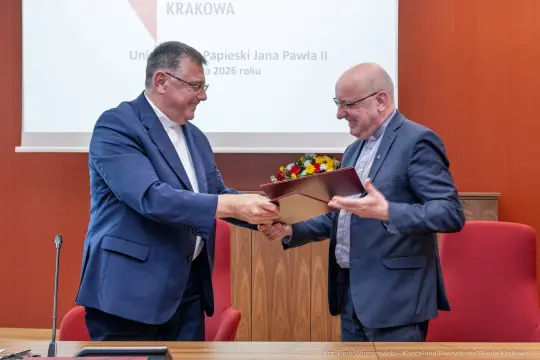 Dwaj mężczyźni wymieniają się dokumentem i kwiatami w sali konferencyjnej.