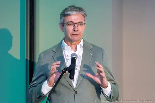 Mężczyzna w szarym garniturze przemawia do mikrofonu na tle zielonej ściany.
