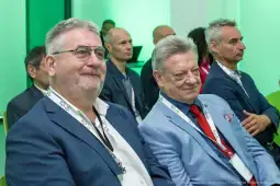 Dwaj mężczyźni w garniturach siedzący na konferencji, w tle inni uczestnicy.
