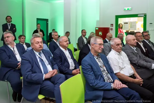 Grupa mężczyzn w garniturach siedzących na zielonych krzesłach podczas konferencji.