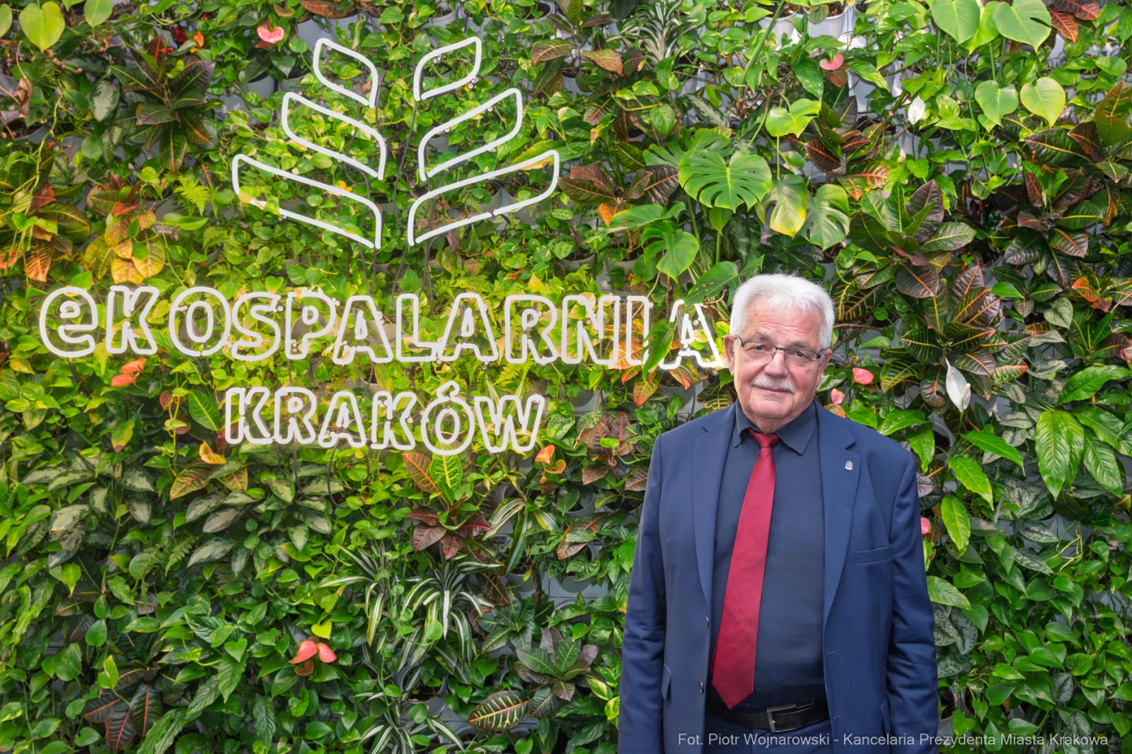 Mężczyzna w garniturze stoi przed zieloną ścianą z napisem Ekospalarnia Kraków.  Autor: Piotr Wojnarowski