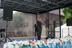 Mężczyzna w garniturze przemawia na scenie, w tle dekoracje kwiatowe.