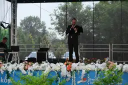 Mężczyzna w garniturze przemawia na scenie, otoczonej kwiatami i zielenią.