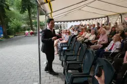 Mężczyzna w garniturze występuje przed grupą starszych osób siedzących na krzesłach.
