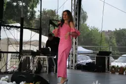 Kobieta w różowej sukience śpiewa na scenie, w tle mężczyzna przy sprzęcie muzycznym.