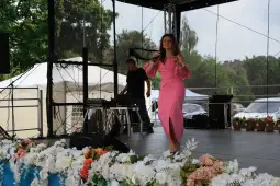 Kobieta w różowej sukni występuje na scenie, w tle mężczyzna przy sprzęcie muzycznym.