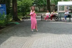 Kobieta w różowej sukience śpiewa na placu, a w tle siedzą widzowie.