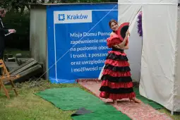 Kobieta w czerwono-czarnym stroju ludowym trzyma wachlarz, pozując na tle banera z napisem o misji Domu Pomocy.