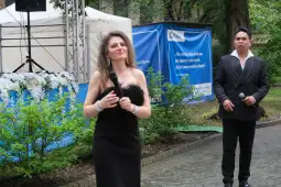 Kobieta w czarnej sukni z mikrofonem, mężczyzna w garniturze z mikrofonem w tle.