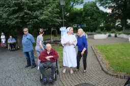 Grupa osób starszych oraz opiekunów w parku, w tym kobieta w białym stroju i mężczyzna na wózku inwalidzkim.