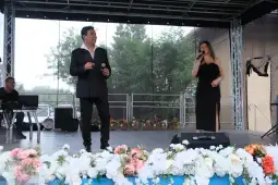 Dwójka artystów występuje na scenie, śpiewając do mikrofonów, w tle kwiaty.