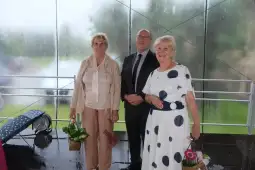 Trzy osoby stojące w deszczu, jedna z kwiatami, dwie w eleganckich strojach.