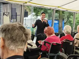 Mężczyzna w garniturze występuje przed grupą osób na wózkach inwalidzkich.