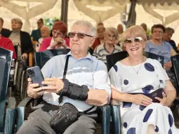Starszy mężczyzna z telefonem i kobieta w białej sukience z niebieskimi kropkami, siedzący w tłumie.