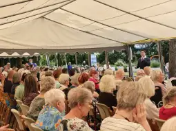 Spotkanie seniorów pod namiotem, z mówcą na scenie i słuchaczami wśród krzeseł.