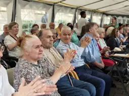Grupa seniorów siedzących w namiocie, niektórzy klaskają, inni obserwują wydarzenie.