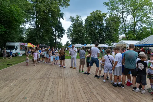 Tłum dzieci i dorosłych na festynie, stoiska z jedzeniem i atrakcjami w tle.
