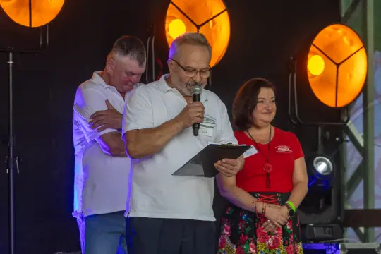 Mężczyzna z mikrofonem przemawia, obok niego dwie osoby, w tle oświetlenie sceniczne.