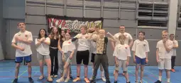 Grupa dzieci i młodzieży w białych koszulkach, pozująca na tle banera z napisem.