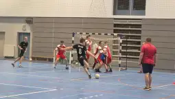 Mecz piłki ręcznej w hali sportowej, zawodnicy w ruchu, bramka w tle.
