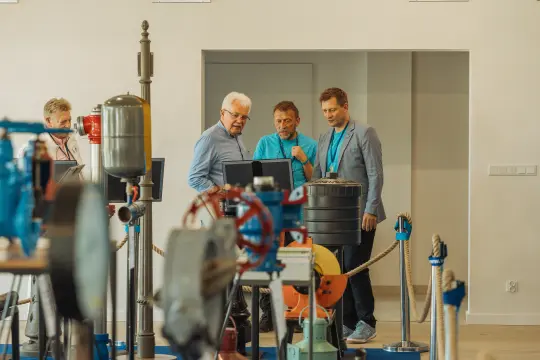 Trzech mężczyzn rozmawia przy komputerze w nowoczesnym pomieszczeniu.