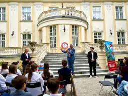 Mężczyzna przemawia na tle eleganckiego budynku, obok dwóch innych mężczyzn i publiczności.