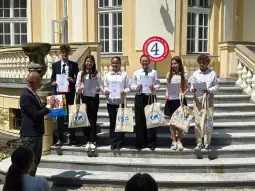 Uczniowie z dyplomami na schodach budynku, obok nauczyciel wręczający nagrody.