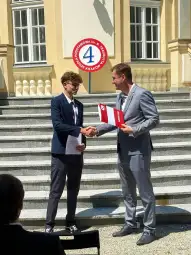 Uroczystość wręczenia dyplomu uczniowi przez nauczyciela na tle budynku.