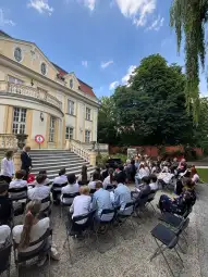 Uczestnicy wydarzenia siedzą na zewnątrz przed budynkiem, w tle drzewa i niebo.