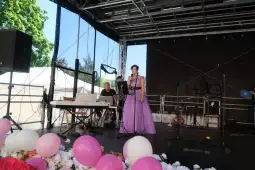 Kobieta w fioletowej sukni śpiewa na scenie, obok muzycy i dekoracje z kwiatów oraz balonów.