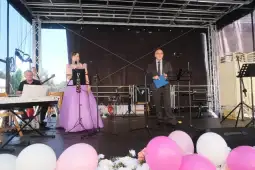 Kobieta w fioletowej sukni i mężczyzna w garniturze przemawiają na scenie, obok muzycy przy instrumentach.