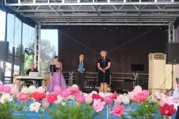Uroczystość na scenie z mówcami i dekoracjami kwiatowymi.