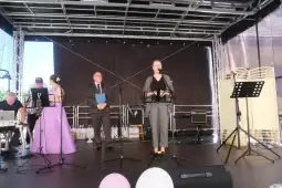 Uroczystość na scenie z mówcami, muzykami i gośćmi w eleganckich strojach.