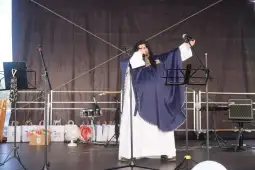 Osoba w kostiumie występuje na scenie, trzyma mikrofon i wykonuje utwór muzyczny.
