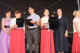 Grupa osób w strojach ludowych podczas występu, trzymających teksty piosenek.