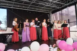 Grupa osób w strojach ludowych śpiewa na scenie, obok instrumenty muzyczne.