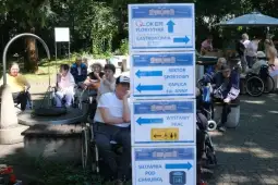 Tablice informacyjne z kierunkami do różnych stref na wydarzeniu, w tle osoby na wózkach inwalidzkich.