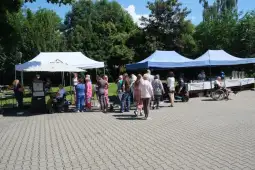 Ludzie w kolejce do stoiska w parku, namioty i zieleń w tle.