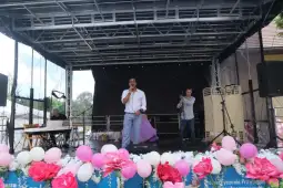 Mężczyzna śpiewa na scenie, obok dwóch innych artystów i dekoracje z kwiatów.