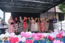 Grupa kobiet w kolorowych sukienkach śpiewa na scenie, otoczone kwiatami i dekoracjami.