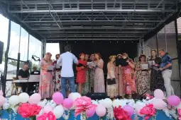 Chór w kolorowych strojach śpiewa na scenie, otoczony kwiatami i balonami.