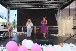 Kobieta w fioletowej sukience i druga w białej bluzce na scenie, obok pianista i dekoracje w różowych balonach.