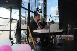 Mężczyzna gra na pianinie, obok dwie osoby siedzą na krzesłach. W tle dekoracje i namioty.