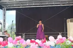 Kobieta w fioletowej sukni śpiewa na scenie, otoczona różowymi balonami i kwiatami.